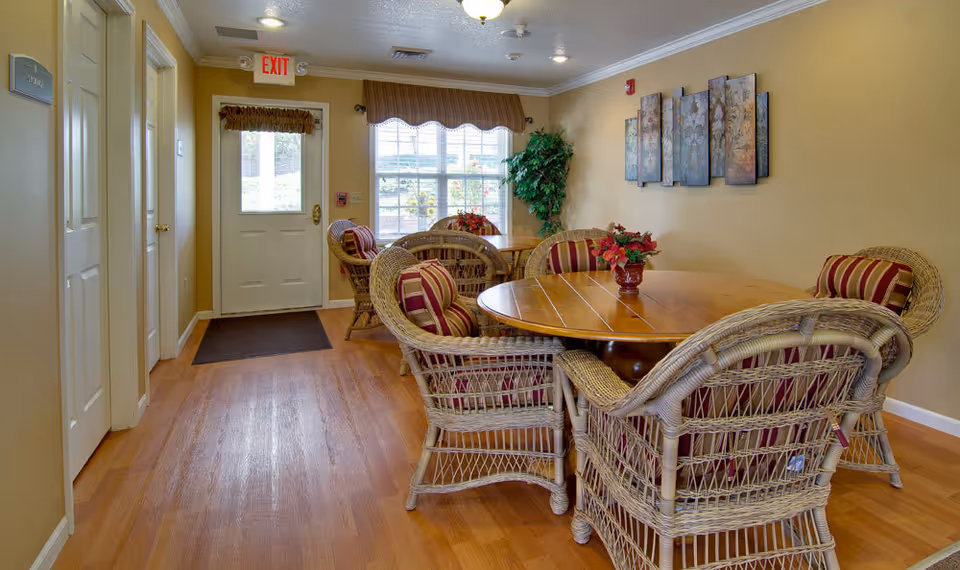 A cozy interior room with wooden flooring featuring two round wooden tables surrounded by wicker chairs with red and beige striped cushions. There are flower arrangements on the tables, a large window with a valance letting in natural light, a potted plant in the corner, and decorative wall art. A white door with an exit sign above it is visible at the far end of the room.