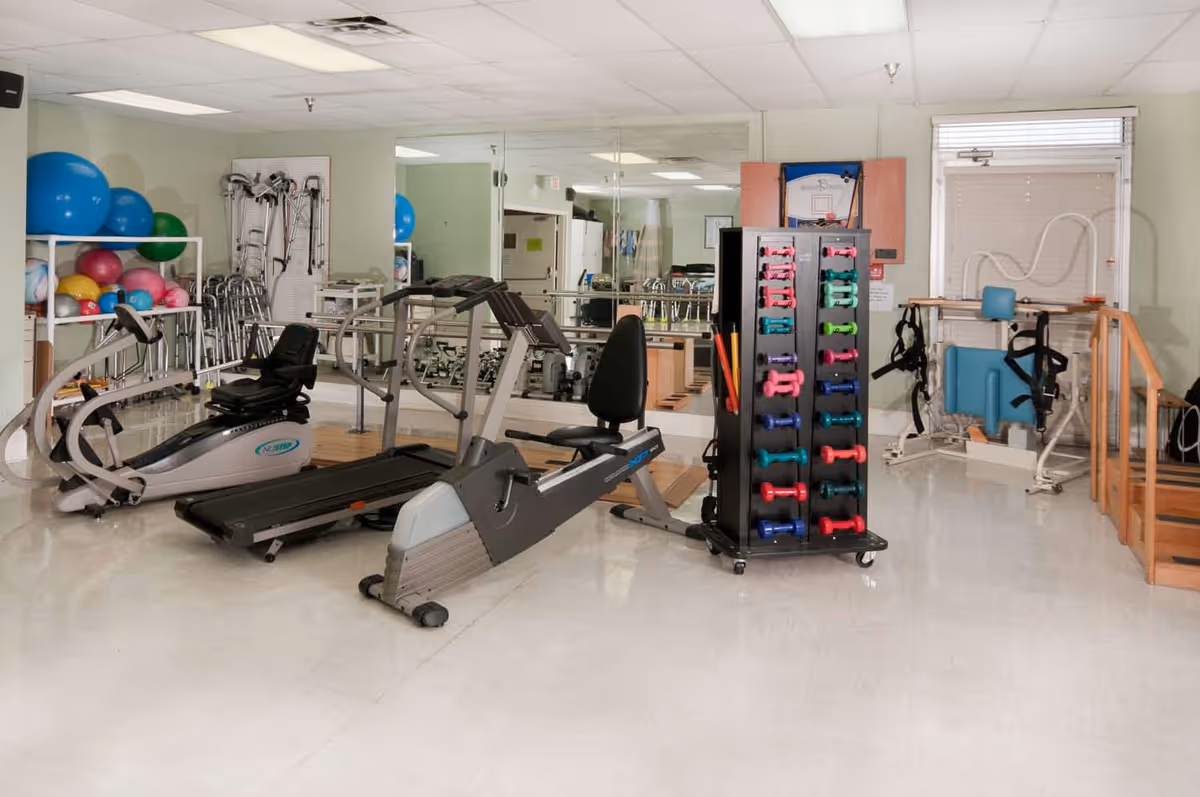 A fitness room in a senior living facility with exercise equipment including a treadmill, stationary bike, a rack of colorful dumbbells, exercise balls, walkers, and other physical therapy equipment. The room has a large mirror on one wall and a window with blinds.