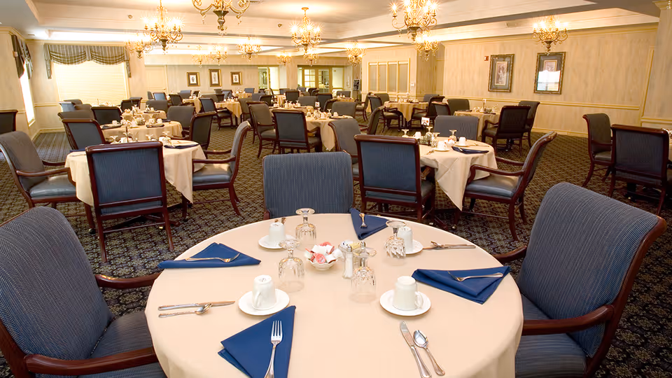 A spacious dining room with multiple round tables covered with beige tablecloths, each set with blue napkins, white cups, glasses, and silverware. The room features patterned carpet, chandeliers, framed artwork on the walls, and large windows with valances allowing natural light.