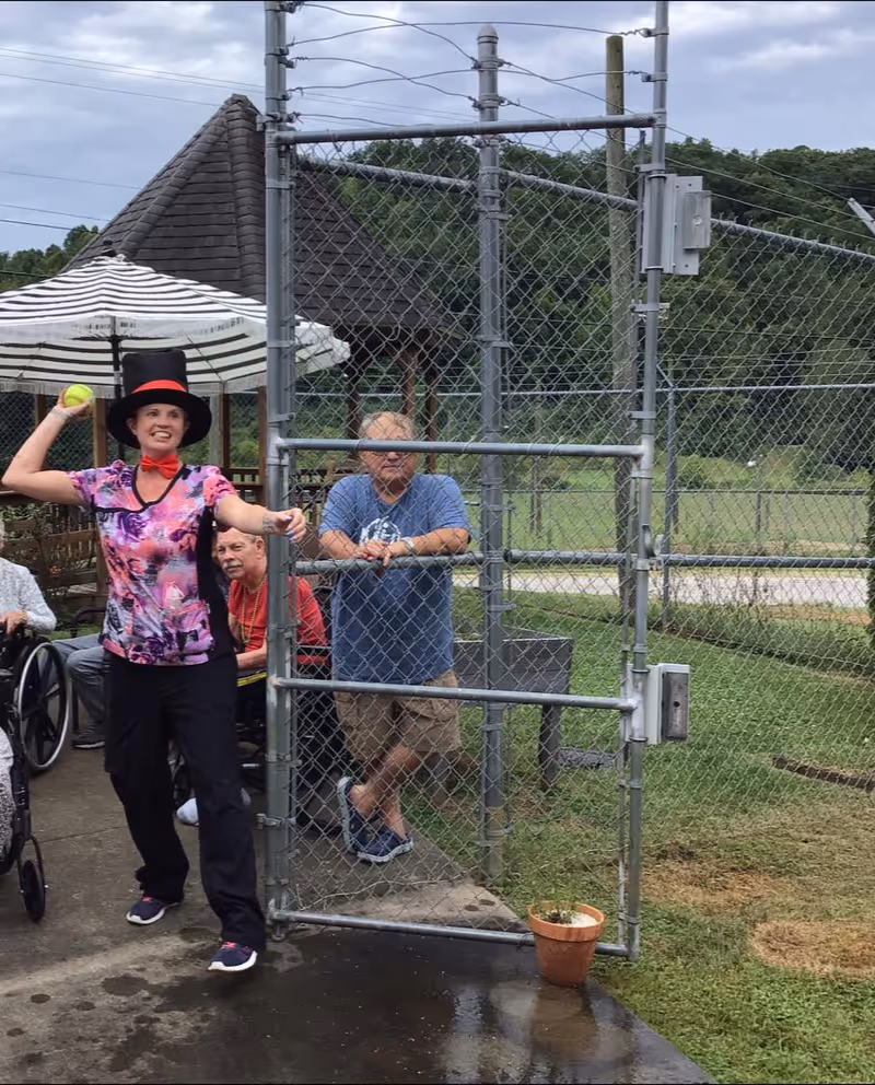 A woman wearing a colorful shirt, black pants, a red bow tie, and a tall black hat is holding a yellow ball and appears to be preparing to throw it. Behind her, there are elderly people sitting and standing near a chain-link fence gate. The setting is outdoors with green grass, trees, and a cloudy sky in the background.