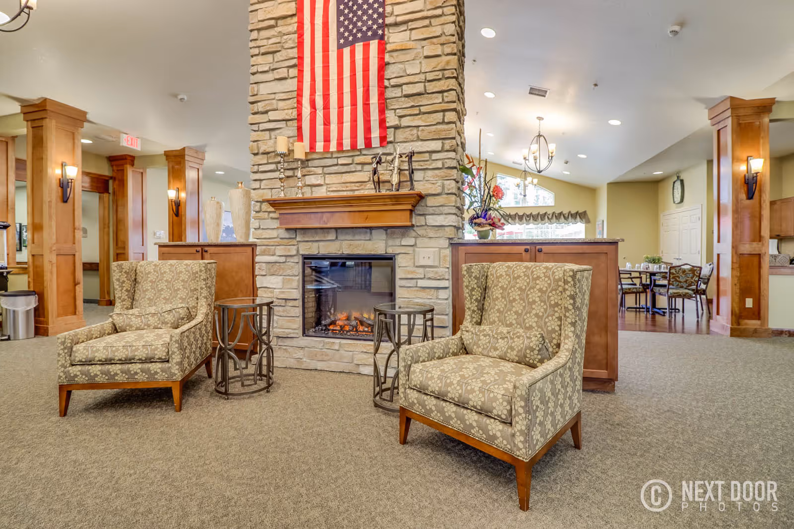 A cozy assisted living facility common area featuring two patterned armchairs facing a stone fireplace with a wooden mantle. An American flag hangs above the fireplace. The room has warm wooden columns, soft lighting, and a dining area with tables and chairs visible in the background.