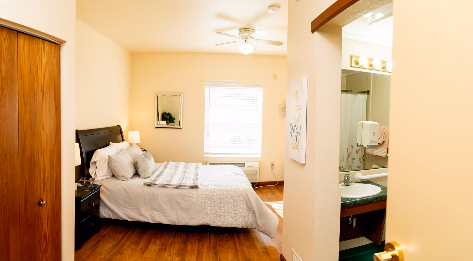 A cozy bedroom with a neatly made bed featuring white and gray bedding, a wooden nightstand with a lamp, a window with blinds, and a ceiling fan. To the right, a partial view of a bathroom sink and mirror is visible through an open doorway.