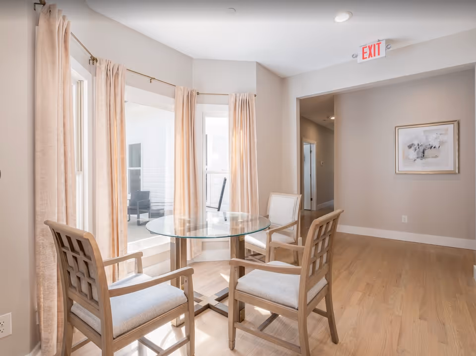 Bright seating area with a round glass table and four wooden chairs by bay windows with beige curtains.
