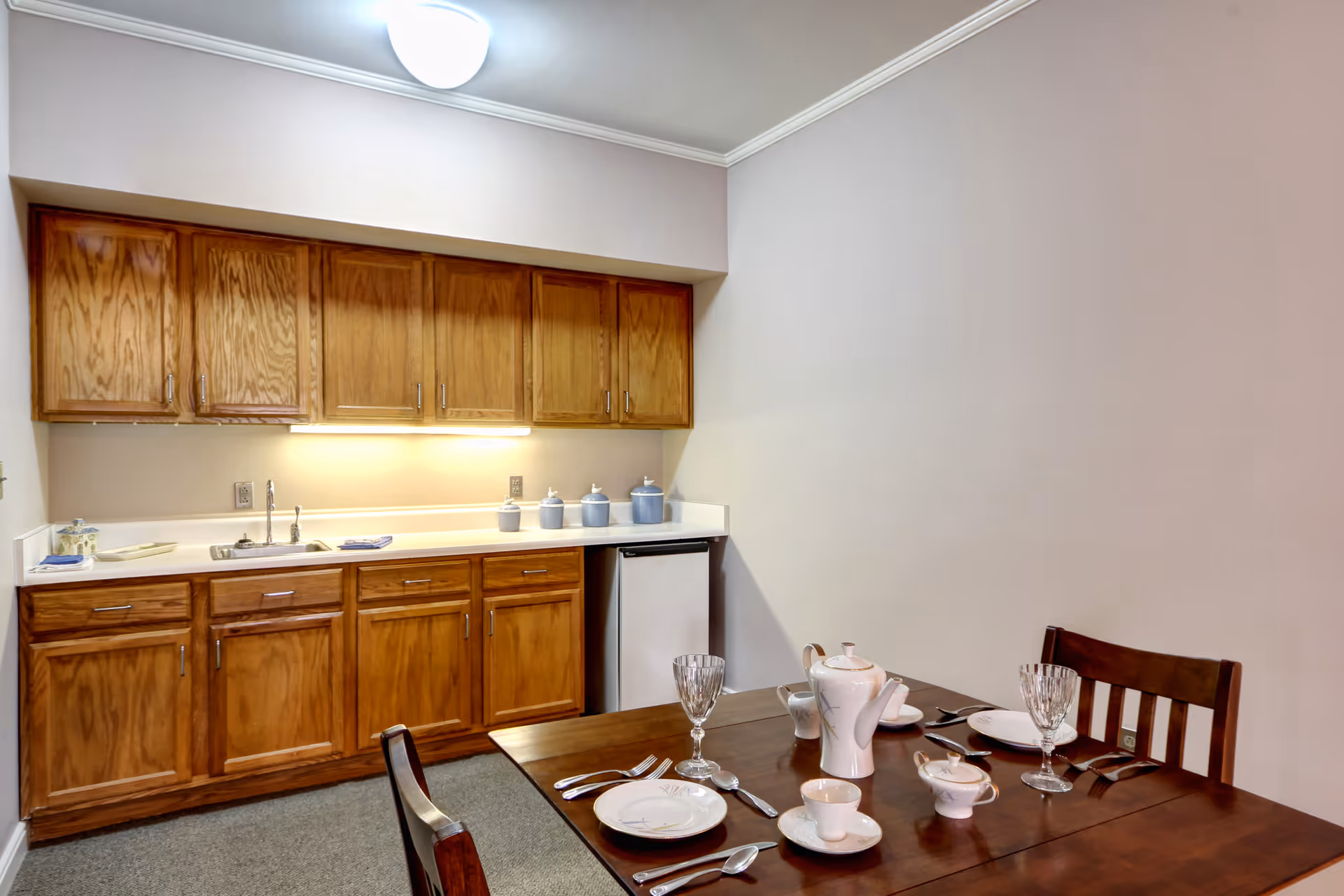 A small kitchen area with wooden cabinets, a sink, a countertop with blue canisters, and a mini refrigerator. In the foreground, there is a wooden dining table set with white china, glassware, and silverware. The walls are plain and light-colored, and the floor is carpeted.