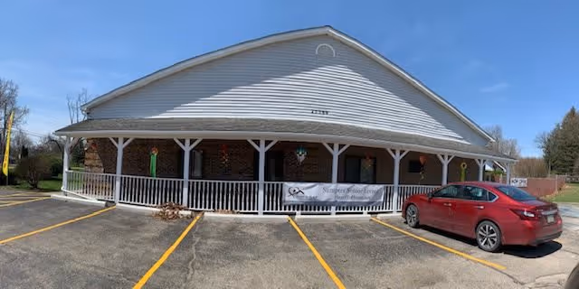 Exterior view of a single-story building with a white roof and brick facade, featuring a covered porch with white railings. A banner on the porch reads 'Sumpter Senior Living'. A red car is parked in one of the parking spaces in front of the building under a clear blue sky.