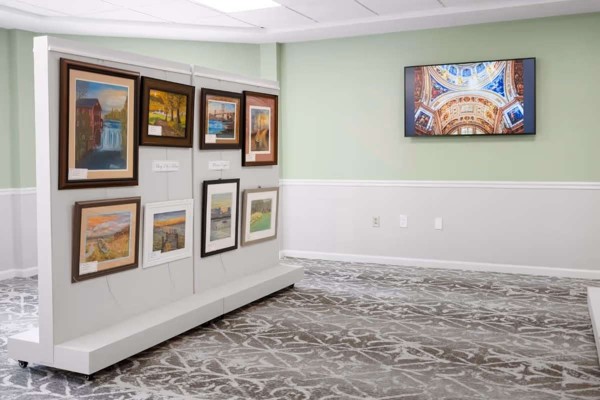 An interior room with light green and white walls, featuring a display panel with framed paintings and artwork. A flat screen TV mounted on the wall shows an image of an ornate ceiling. The floor is covered with a patterned carpet.