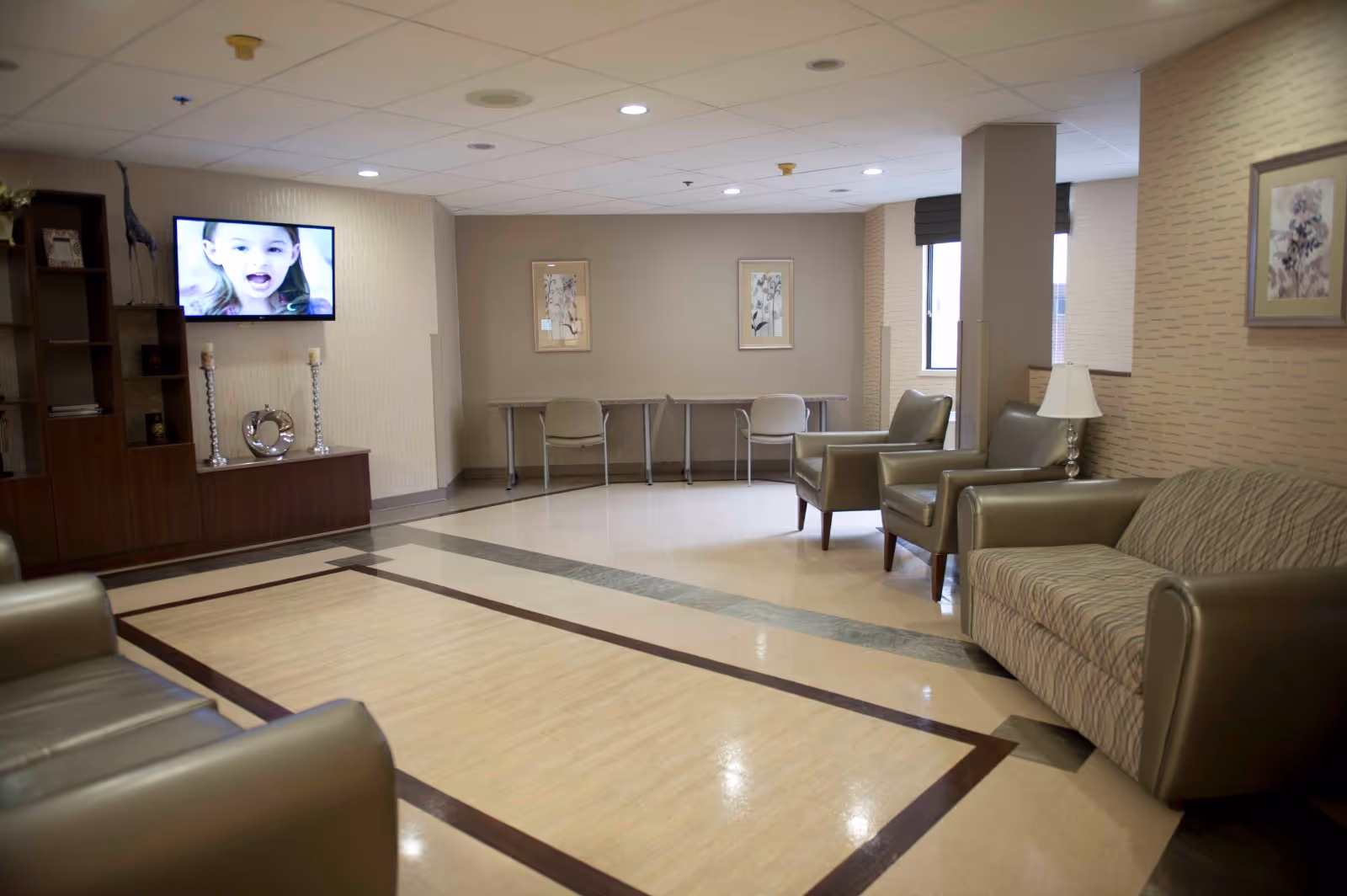 A well-lit living room area in a healthcare facility with beige walls and flooring. The room features several cushioned chairs and a sofa arranged around the perimeter. There is a flat-screen TV mounted on the wall displaying a child's face, a wooden cabinet with decorative items, two framed pictures on the far wall, and a window with blinds letting in natural light.
