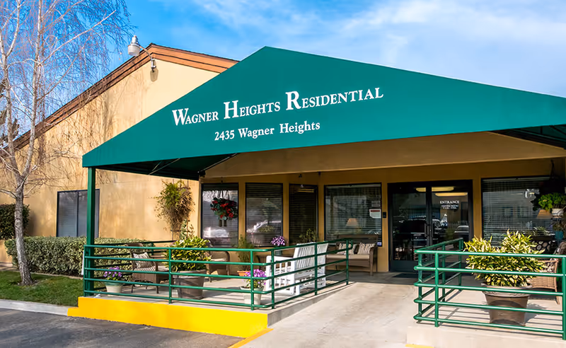 Entrance of Wagner Heights Residential facility with a green awning displaying the facility name and address. The entrance area has a ramp with green railings, potted plants, and outdoor seating including chairs and a bench. The building exterior is beige with windows and a glass door labeled 'Entrance'.