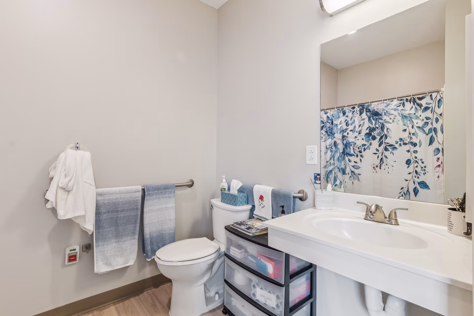 A clean bathroom with a white toilet, a white sink with a silver faucet, and a large mirror above the sink. There are two blue and white towels hanging on a towel rack next to the toilet, and a white towel hanging on a hook on the wall. A shower curtain with a blue and green leafy pattern is visible in the mirror. A small storage unit with drawers is placed next to the toilet, holding various items including tissues and hand soap.