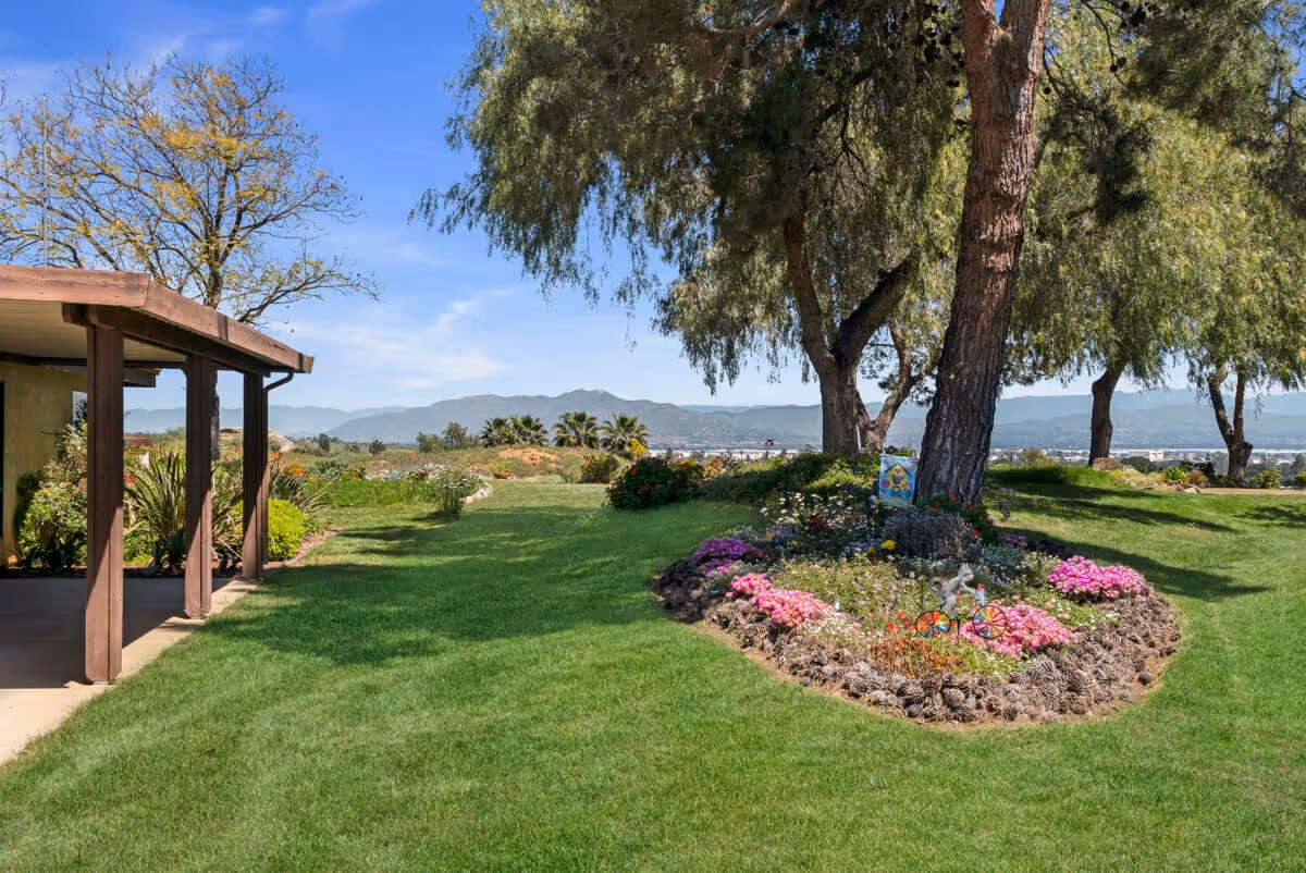 A well-maintained outdoor garden area with green grass, colorful flower beds, and several large trees providing shade. There is a covered patio structure on the left side and mountains visible in the distance under a clear blue sky.
