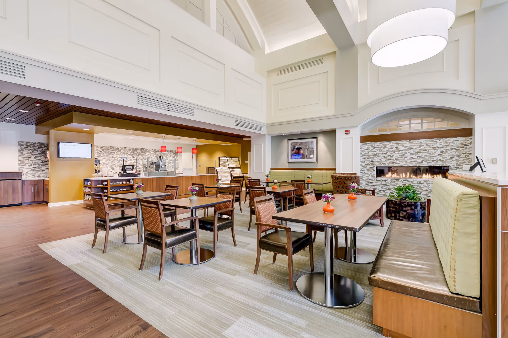 Bright and spacious dining area in Copper Ridge facility featuring multiple wooden tables with chairs and booth seating. The room has a modern design with a fireplace set in a tiled wall, a high ceiling with exposed beams, and pendant lights above the serving counter in the background.