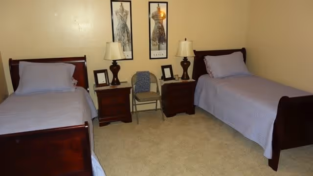 A bedroom with two single wooden beds, each with a light purple bedspread and pillow. Between the beds are two wooden nightstands, each with a lamp and framed pictures. A folding chair with a folded blanket is placed between the nightstands. Two framed fashion sketches hang on the beige wall above the nightstands.