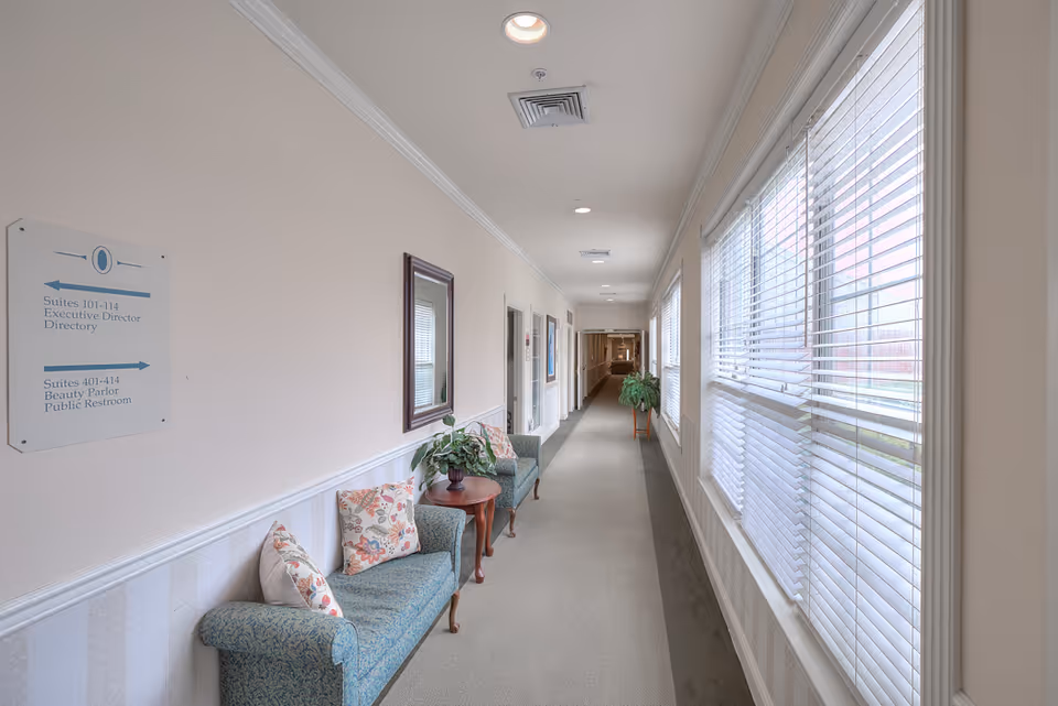 A long, well-lit hallway in a senior living facility with large windows on the right side covered by white blinds. On the left side, there are two blue upholstered chairs with floral cushions, a small wooden table with a potted plant, a wall mirror, and a directory sign indicating suite numbers and locations. The hallway has beige walls with white wainscoting and a carpeted floor.