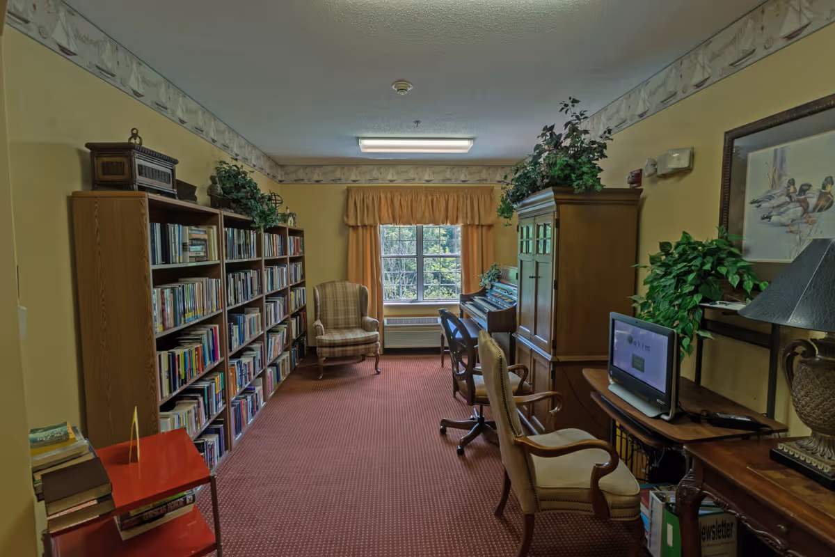 Cozy communal library room with tall bookshelves along the left wall, armchairs, a piano, and a desk with a computer.