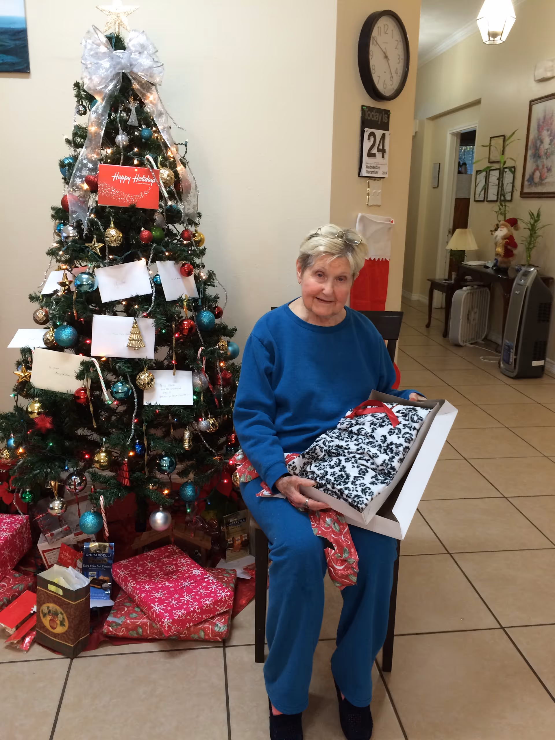 An elderly woman sits beside a decorated Christmas tree in a communal room holding a wrapped gift.