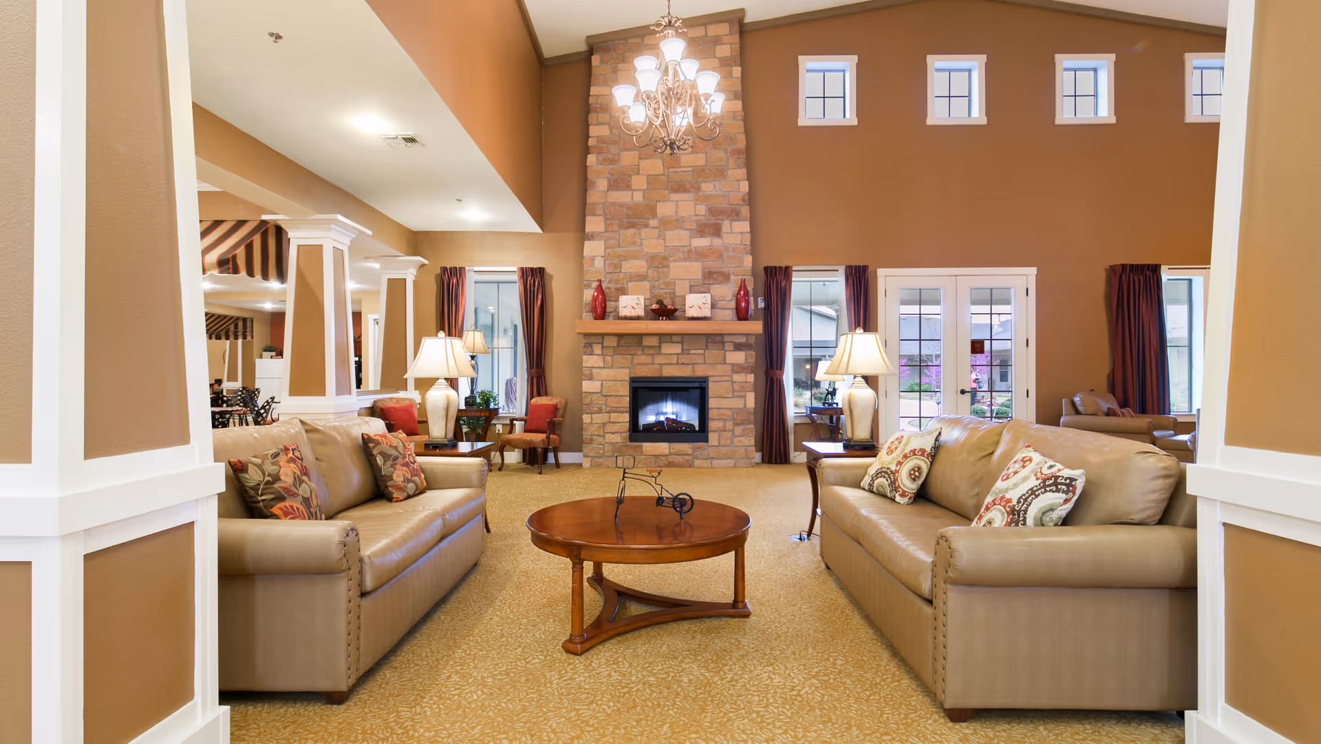 A spacious living room with two beige leather sofas facing each other, a round wooden coffee table in the center, and a stone fireplace with a lit fire in the background. The room has high ceilings with a chandelier, several windows with dark curtains, and warm brown walls. There are also table lamps on side tables next to the sofas and additional seating visible in the background.