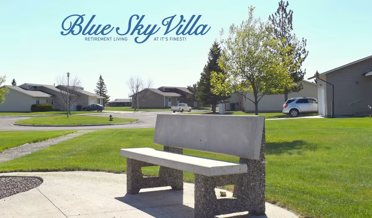 Outdoor scene at Blue Sky Villa Retirement Village showing a concrete bench on a paved area surrounded by green grass, trees, and several single-story residential buildings under a clear blue sky.