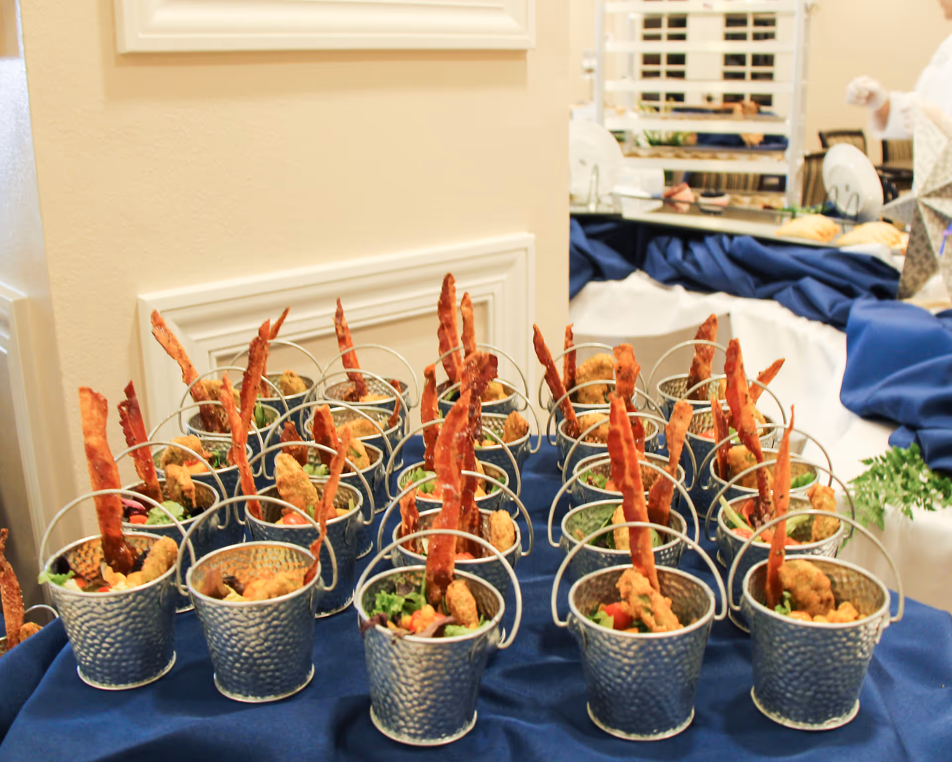 Small hammered metal buckets filled with salad greens, fried chicken strips, and crispy bacon strips arranged on a blue tablecloth in an indoor setting.