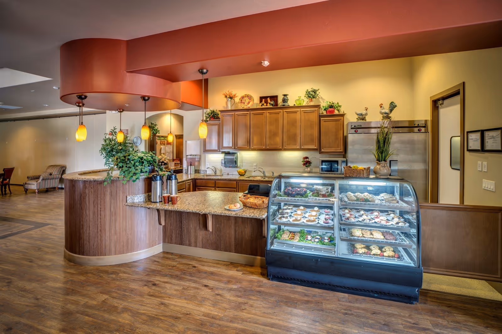 Interior view of a senior living facility's food service area with a curved wooden counter, granite countertop, hanging pendant lights, and a glass display case filled with various pastries and desserts. Wooden cabinets and a stainless steel refrigerator are visible in the background, along with decorative plants and kitchen appliances.