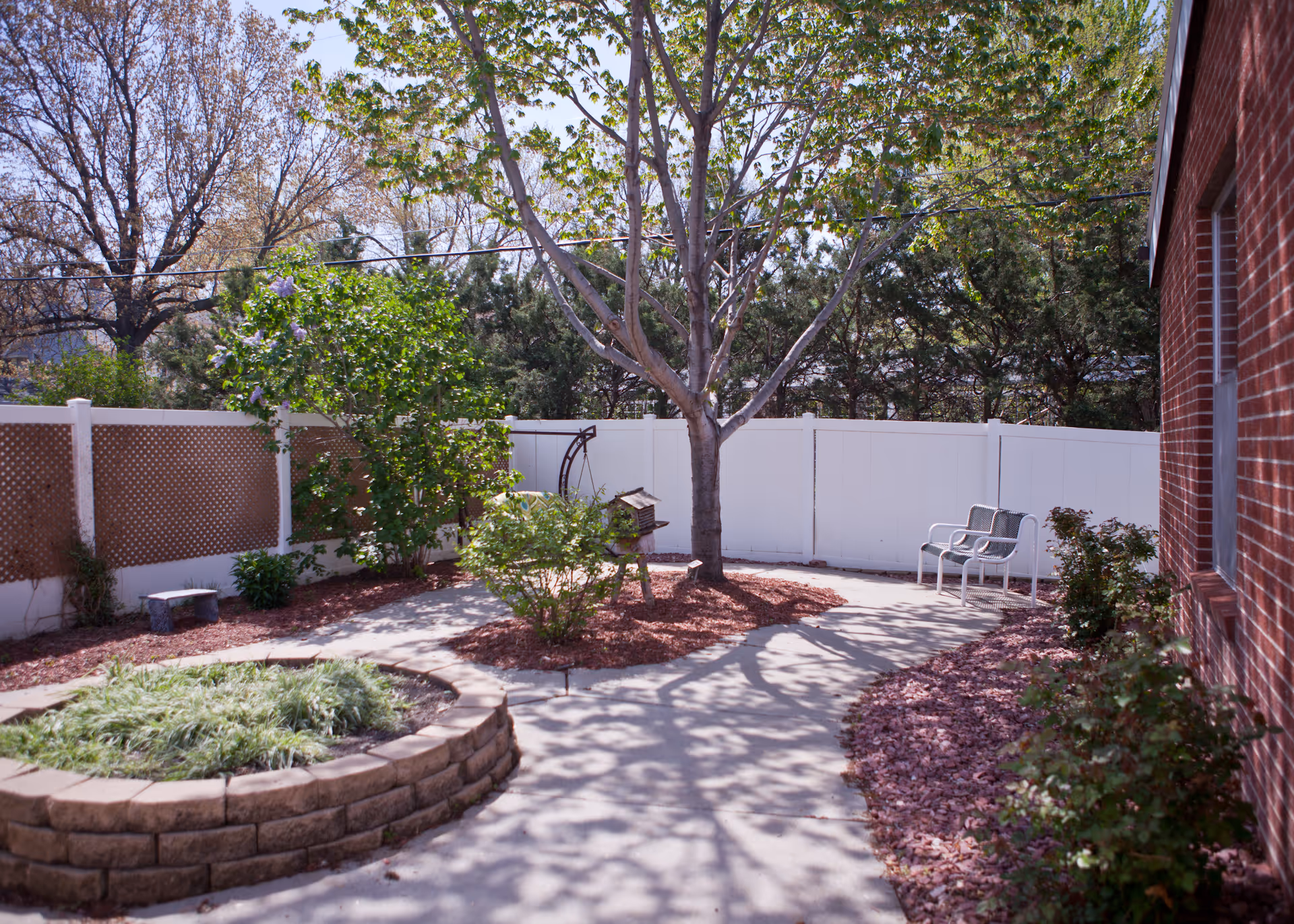 Outdoor garden area with a concrete pathway, a tree in the center, green shrubs, and two metal chairs near a white fence. The area is bordered by a brick building on the right and has a raised circular planter with greenery on the left.