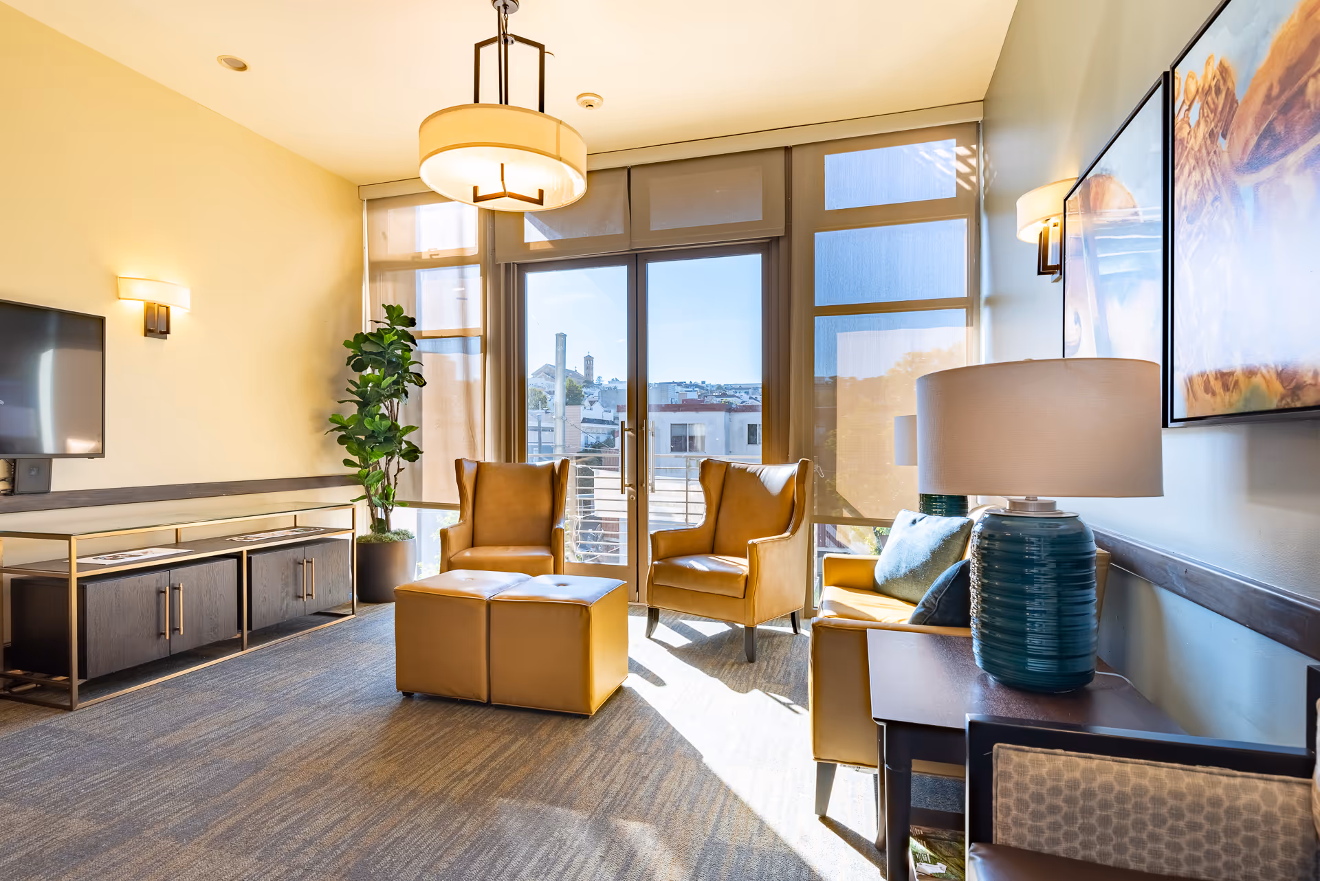 Bright sunlit living room with two leather armchairs, an ottoman, TV console, and large floor-to-ceiling windows and door.