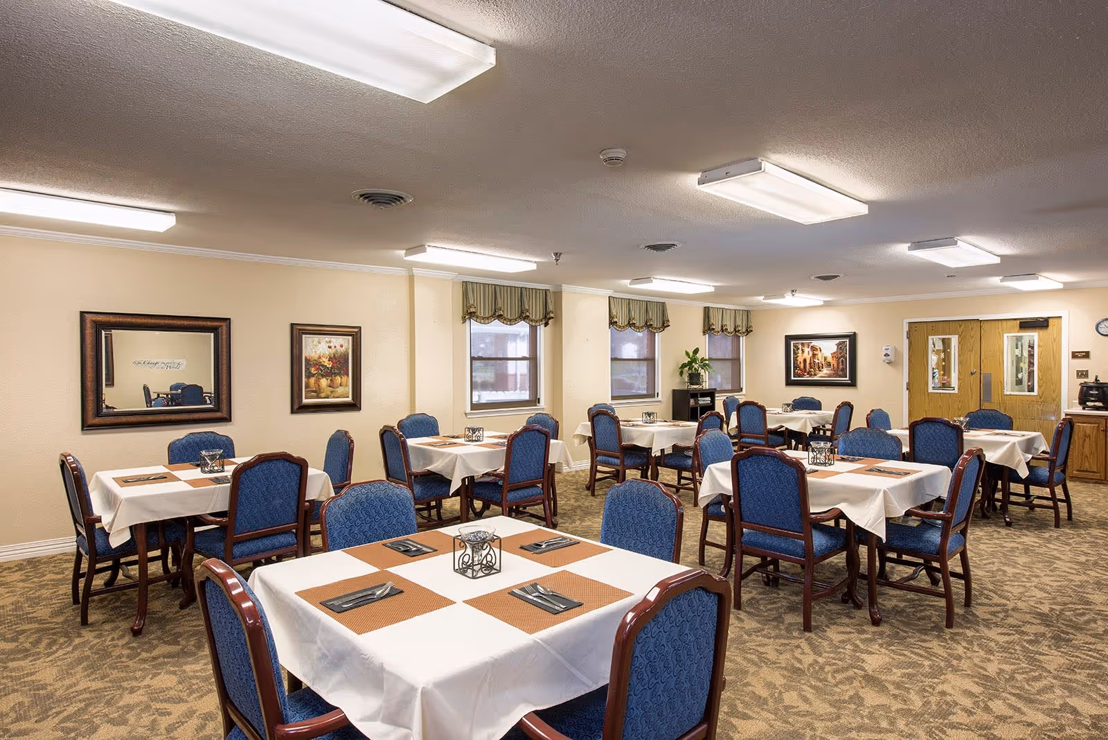 Dining room with several square tables set with placemats and blue upholstered chairs.