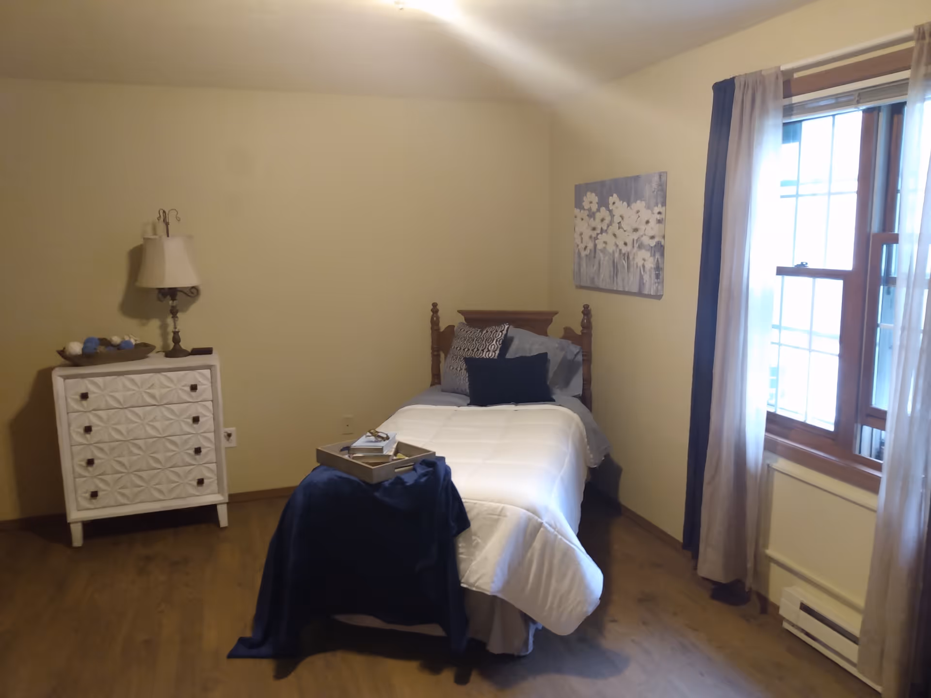 A small bedroom with a single bed covered in a white comforter and multiple pillows. There is a small white dresser with a decorative lamp and a bowl of decorative balls on top. A tray with books and glasses is placed on a blue cloth at the foot of the bed. The room has wooden flooring, beige walls, a window with curtains, and a floral painting on the wall.