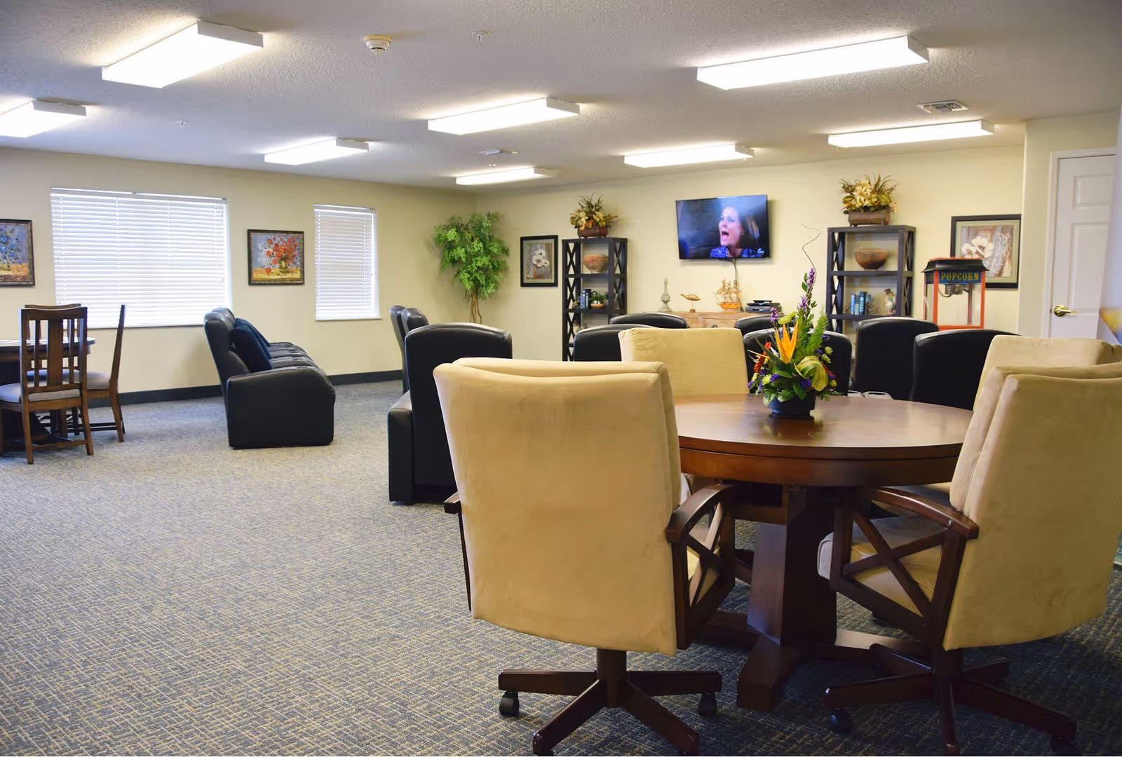 A spacious senior living common area with a round wooden table surrounded by beige cushioned chairs in the foreground. In the background, there are black leather sofas, a flat-screen TV mounted on the wall, decorative plants, framed artwork, and shelves with decorative items. The room is well-lit with ceiling lights and has carpeted flooring.