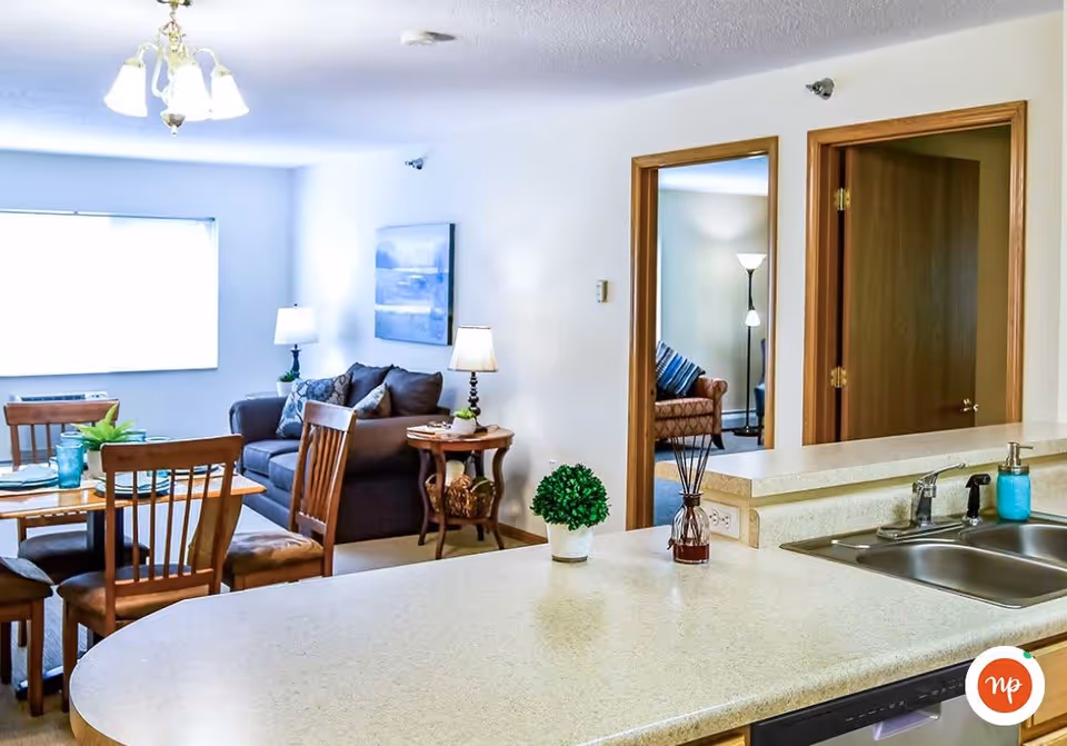 View of a senior living facility interior showing a kitchen counter with a sink and soap dispenser in the foreground, a dining table with chairs, a living room area with a sofa, side table with a lamp, and two doorways leading to other rooms with additional seating and lamps.