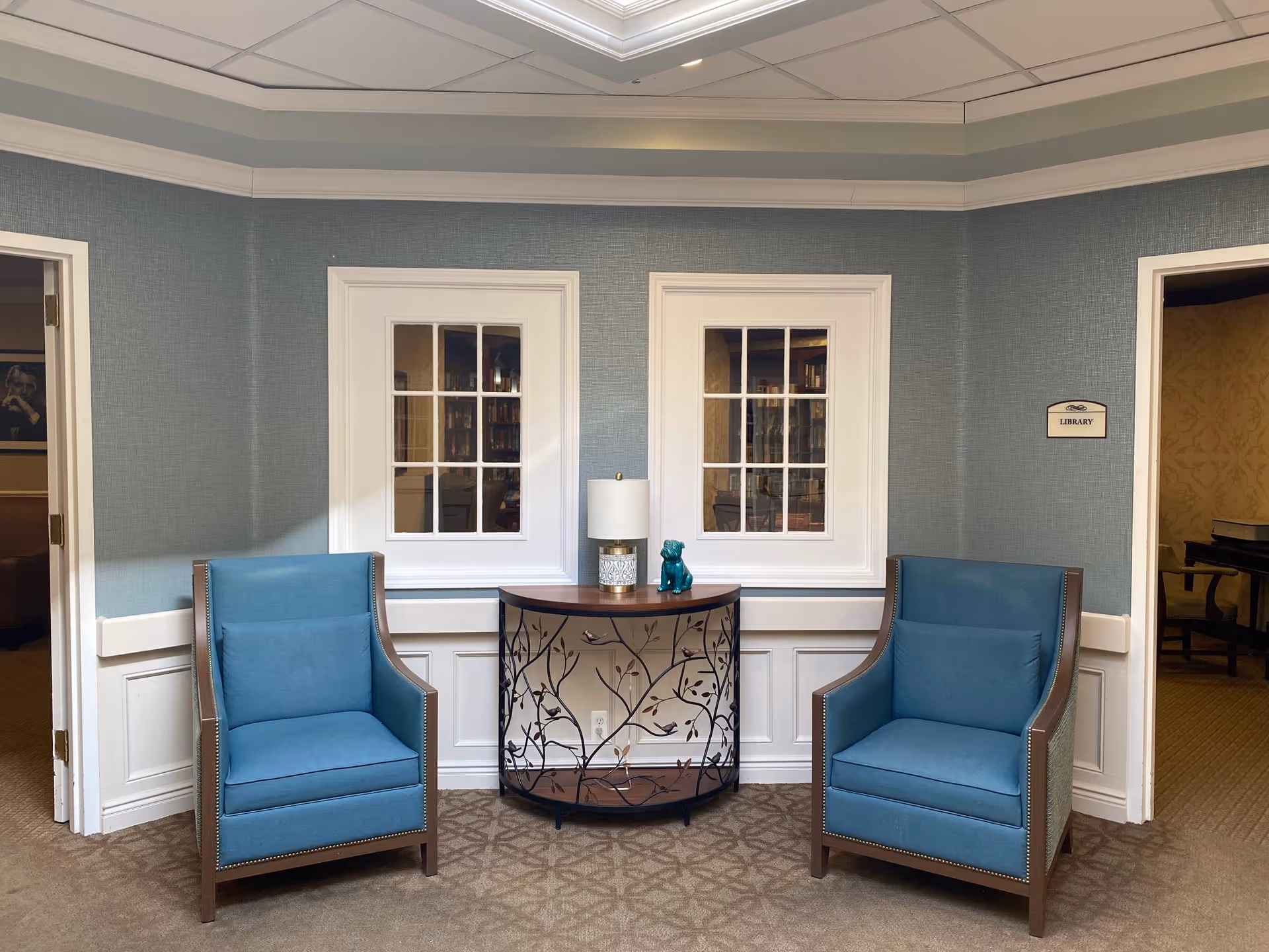Two blue armchairs flank a decorative console table with a lamp and figurine in a hallway seating area outside the library.