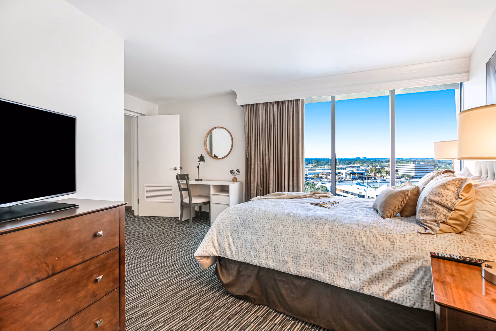 A bright bedroom with a large bed covered in patterned bedding and multiple pillows. There is a wooden dresser with a flat-screen TV on top to the left, and a small desk with a chair and a round mirror on the wall in the background. Large windows with beige curtains offer a view of a cityscape outside.