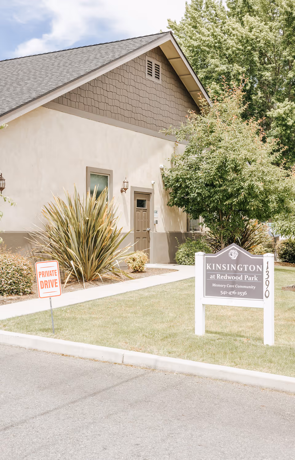 Exterior view of a building at Kinsington at Redwood Park, a memory care community. The building has beige walls, a brown door, and is surrounded by greenery including bushes and trees. There is a sign in front of the building displaying the community name and contact number, and a smaller sign indicating a private drive.