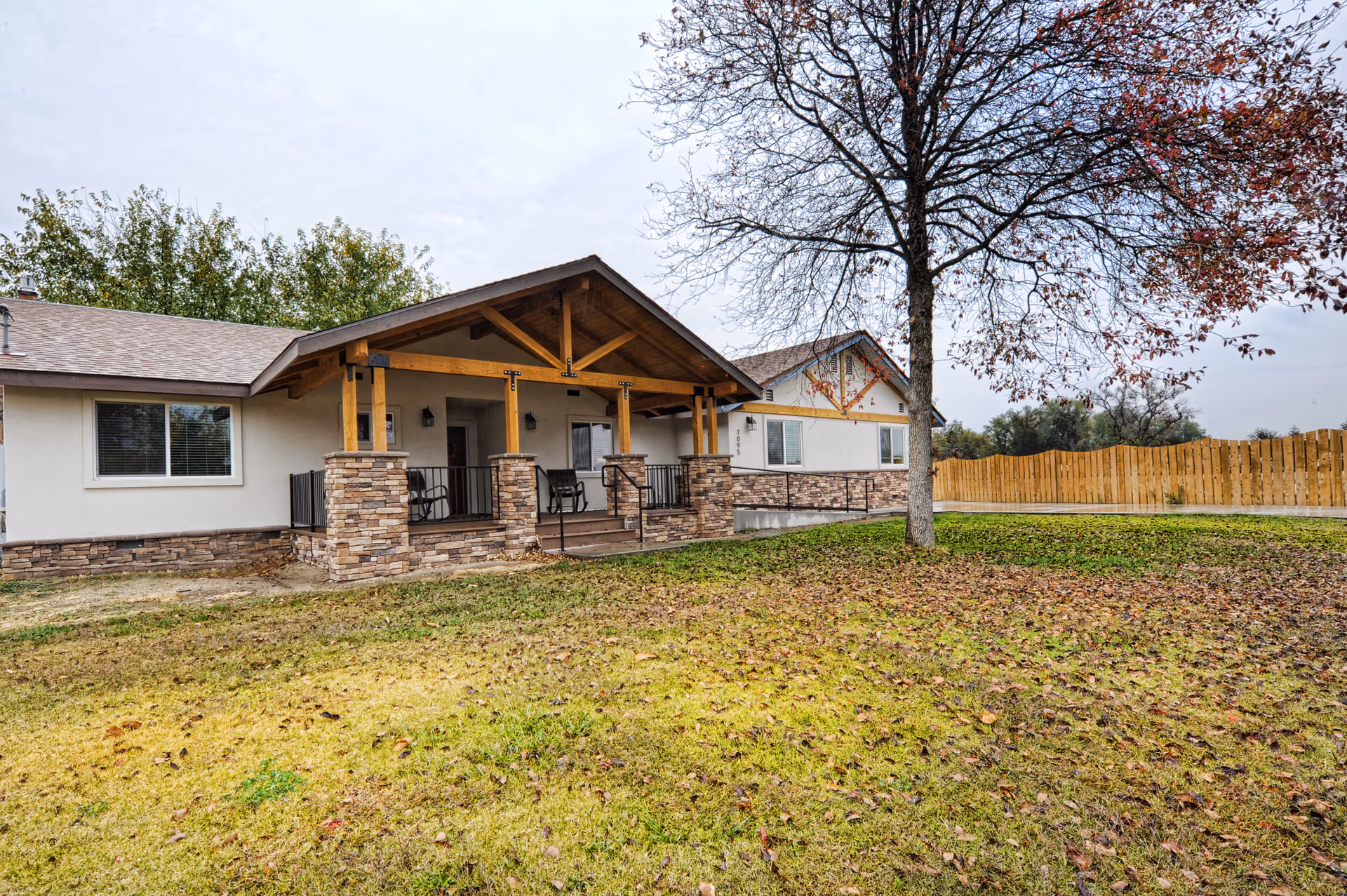 Exterior view of a single-story assisted living facility building with a covered porch supported by wooden beams and stone pillars. The building is surrounded by a grassy yard with a tree in the foreground and a wooden fence in the background under a cloudy sky.