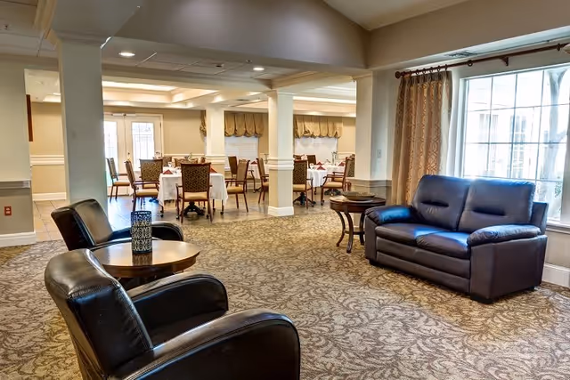 A spacious common area in a senior living facility featuring two black leather armchairs and a black leather sofa arranged around wooden side tables. In the background, there is a dining area with multiple tables covered with white tablecloths and chairs. Large windows with curtains allow natural light to fill the room.