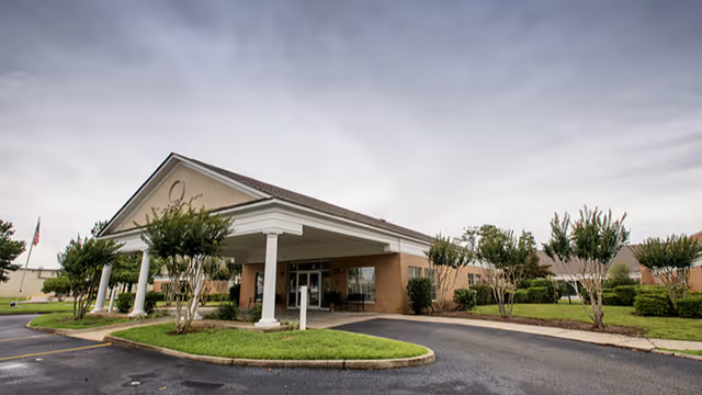 Exterior view of a single-story brick building with a covered entrance supported by white columns, surrounded by trimmed bushes and small trees under a cloudy sky.