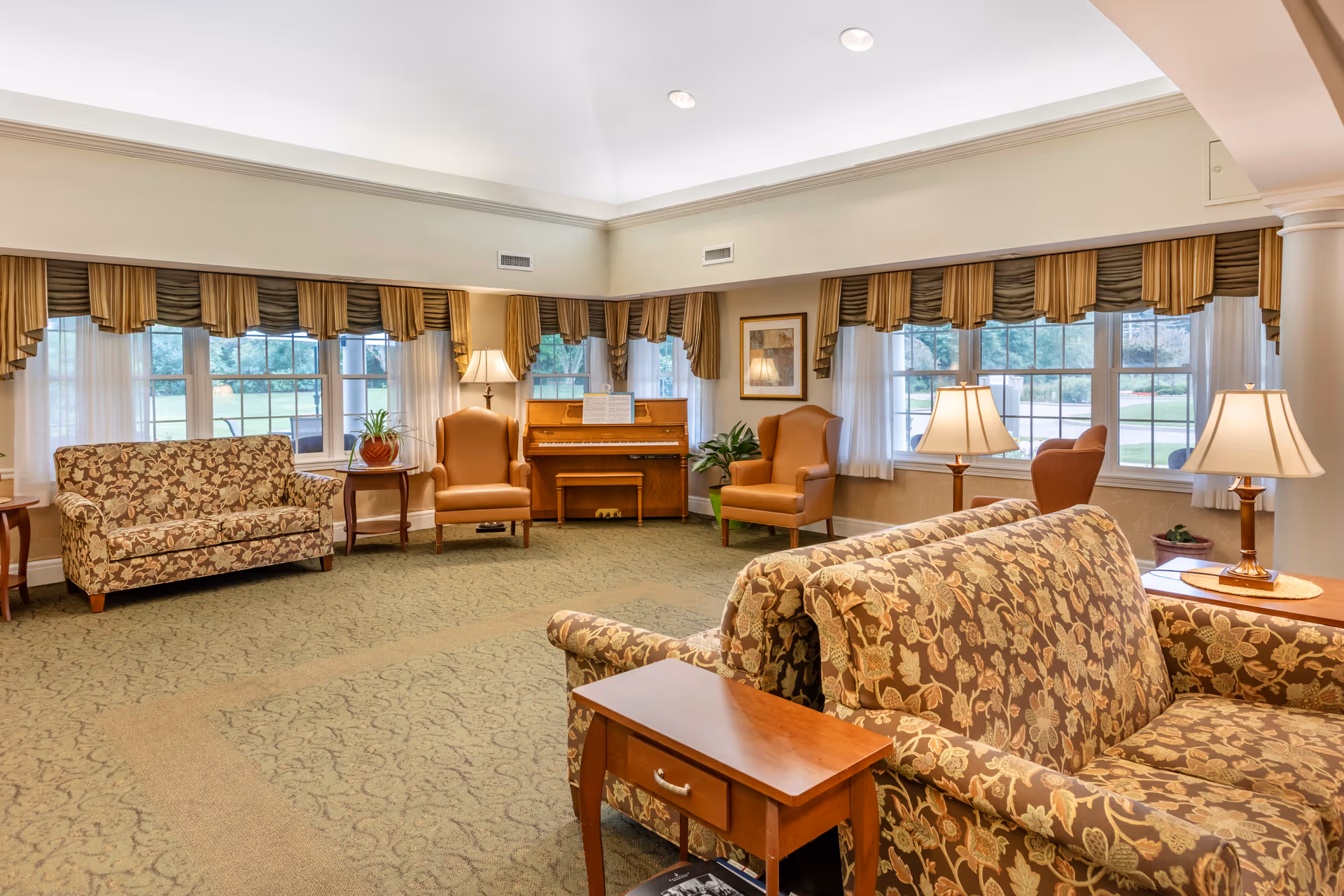 A spacious living room area with floral patterned sofas, two brown armchairs, wooden side tables with lamps, a piano against the wall, large windows with valance curtains, and a green carpeted floor.