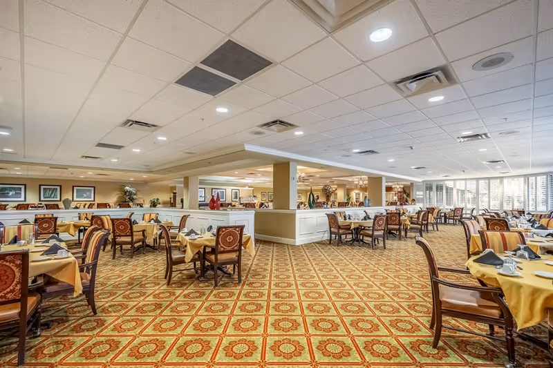 Large formal dining room with many tables set with yellow tablecloths, upholstered chairs, and patterned carpet.
