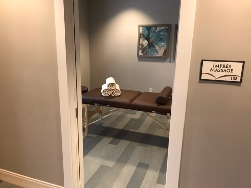 View through an open doorway into a massage room with a brown massage table. Three rolled white towels are stacked on the table. A framed floral artwork hangs on the gray wall in the background. A sign on the wall outside the room reads 'IMPRÉS MASSAGE 108'.