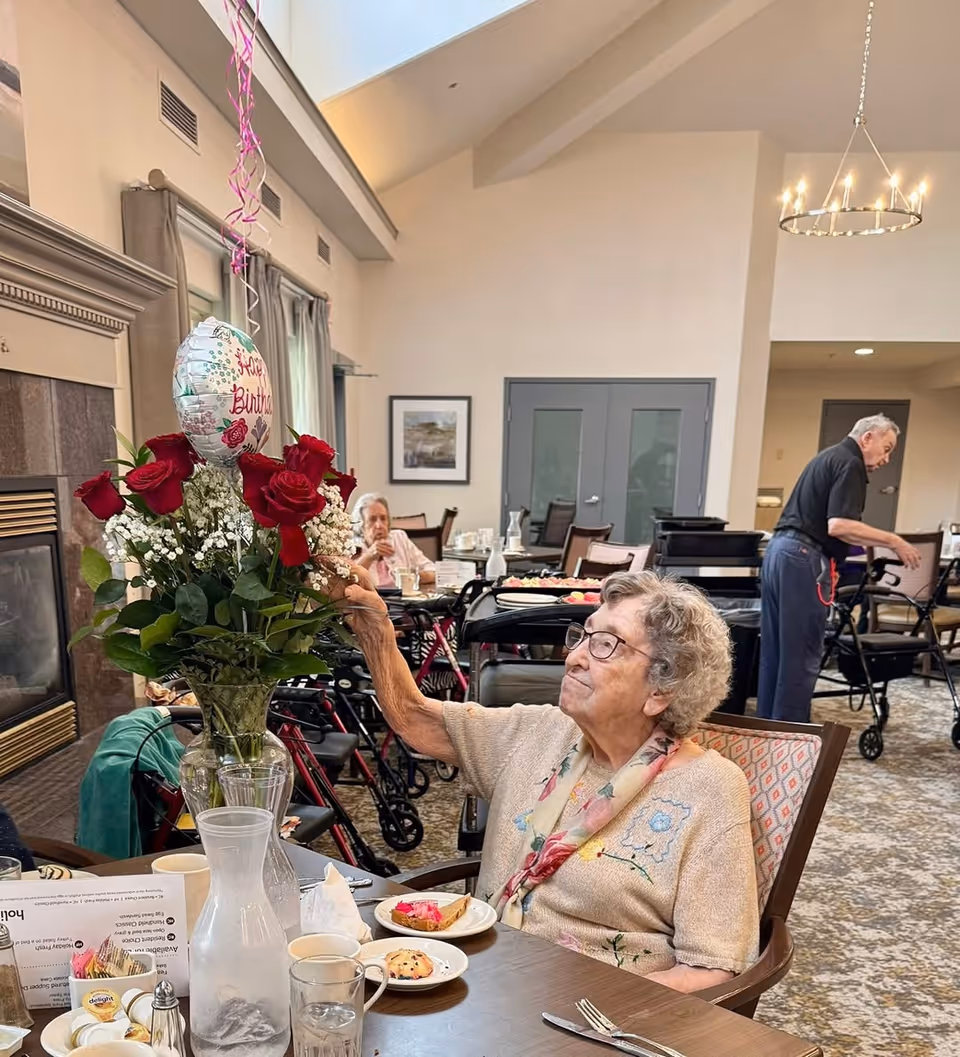 An elderly woman sitting at a dining table in a senior living facility, reaching to touch a bouquet of red roses with a 'Happy Birthday' balloon attached. The table has plates with food, cups, and a water pitcher. In the background, other elderly residents are seated or walking with walkers in a spacious, well-lit room with high ceilings and a chandelier.