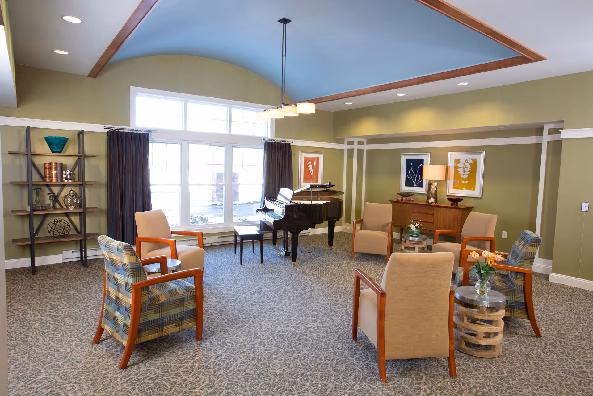 A spacious and well-lit living room with a grand piano near large windows, surrounded by six comfortable armchairs arranged in a circle. The room features green walls, framed artwork, a wooden sideboard with a lamp, and decorative shelves with various items. The ceiling has a blue inset with wooden trim and a modern light fixture.