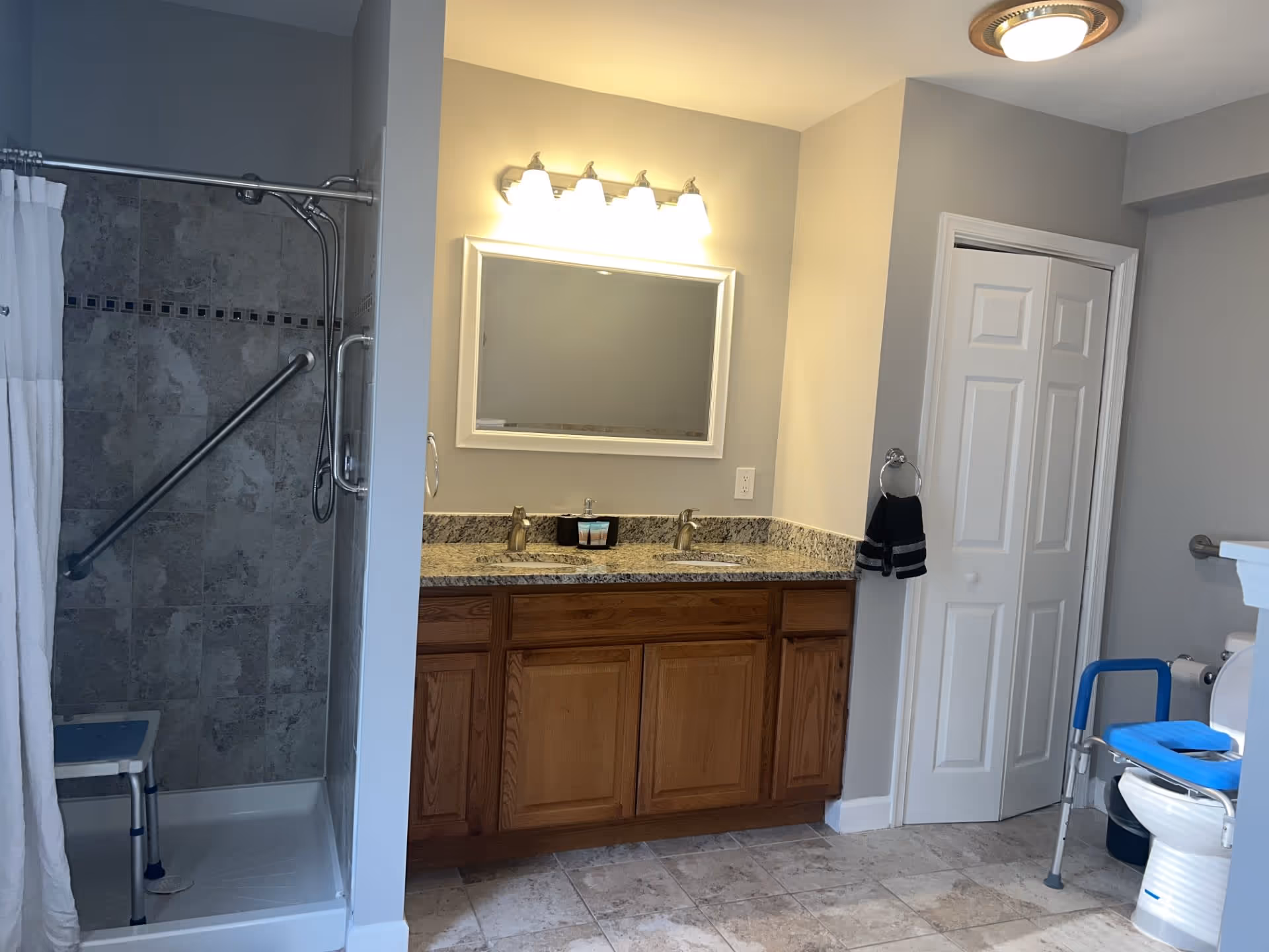 A bathroom with a tiled walk-in shower featuring a grab bar and a shower chair, a double sink vanity with granite countertop and wooden cabinets, a large mirror with four lights above it, a white door closet, and a toilet equipped with a blue raised seat and grab bars.