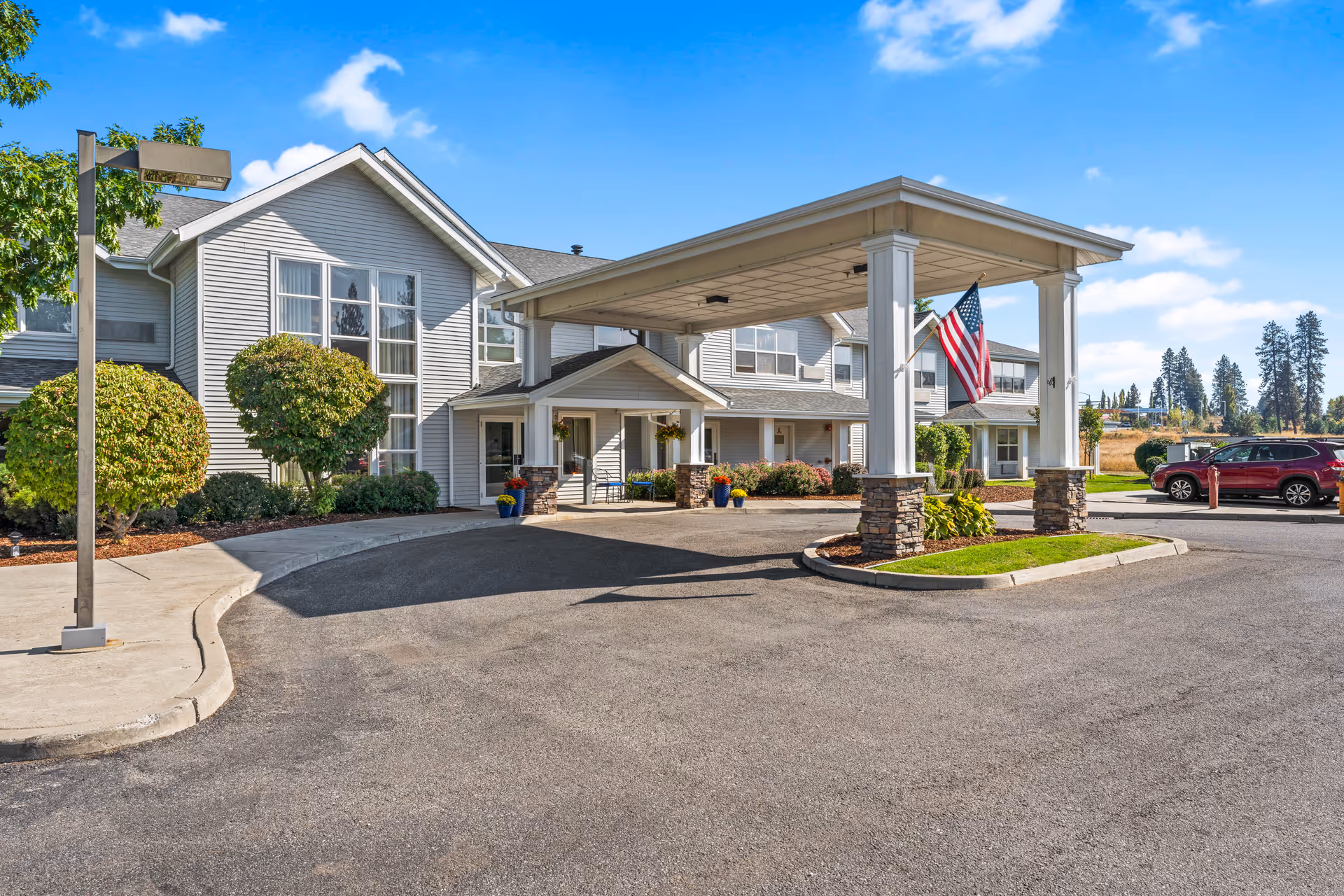 Exterior view of Legends Park Assisted Living facility showing a large covered entrance with stone pillars and an American flag. The building is two stories with gray siding and large windows. There are neatly trimmed bushes and a paved driveway with parked cars under a clear blue sky.