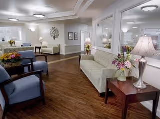 Bright furnished common lounge with sofas, armchairs, side tables, lamps, and floral arrangements.