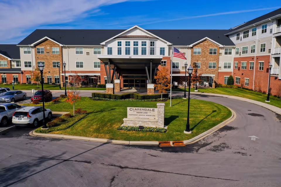 Front exterior view of Clarendale at Indian Lake, a multi-story senior living facility with brick and siding facade, a covered entrance, an American flag, landscaped lawn, and a parking area with several cars.