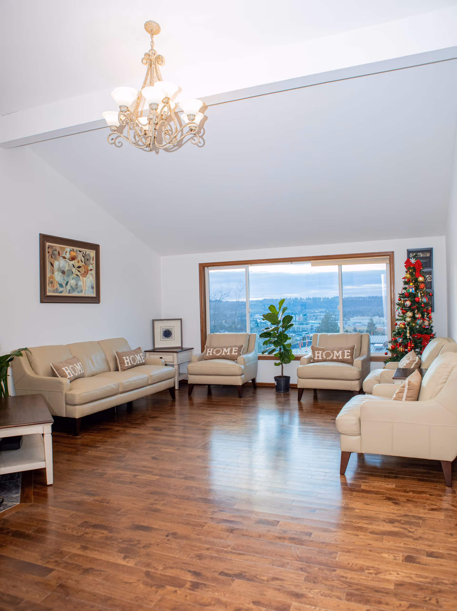 A spacious living room with wooden flooring, beige leather sofas and armchairs with 'HOME' pillows, a large window showing an outdoor view, a decorated Christmas tree in the corner, a chandelier hanging from the ceiling, and framed artwork on the wall.