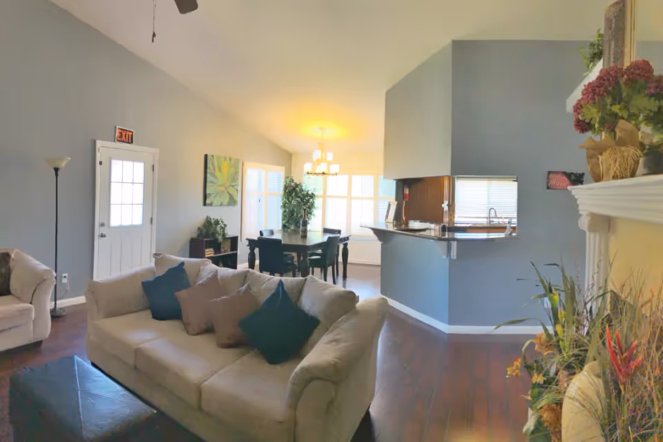 A cozy living room area with beige sofas adorned with dark and light throw pillows, a dark wooden coffee table, and a floor lamp near a white door with an exit sign above it. The room features a dining area with a dark wooden table and chairs, a large window letting in natural light, and a kitchen with a breakfast bar and sink. Decorative plants and flowers are placed around the room, including on a white mantelpiece.
