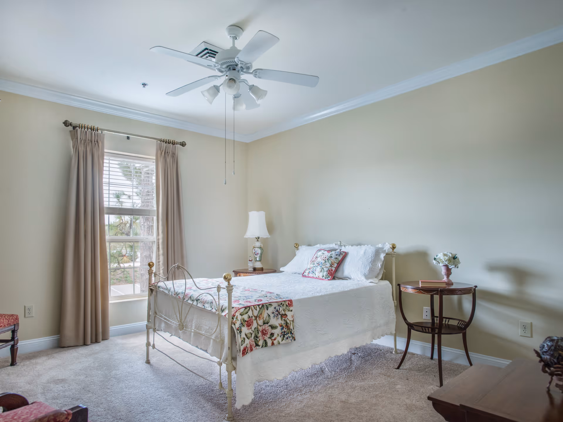 A bright bedroom with a white metal bed frame, a white bedspread, and floral pillows and a floral throw at the foot of the bed. There is a window with beige curtains on the left wall, a ceiling fan with lights above the bed, a wooden side table with a lamp on the left side of the bed, and a round wooden side table with a small flower arrangement on the right side. The room has beige walls and carpeted flooring.