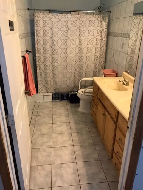 A bathroom with tiled floor and walls, featuring a shower with a patterned curtain, a toilet with a safety grab bar, a wooden vanity with a sink, and a pink towel hanging on a wall-mounted towel rack.