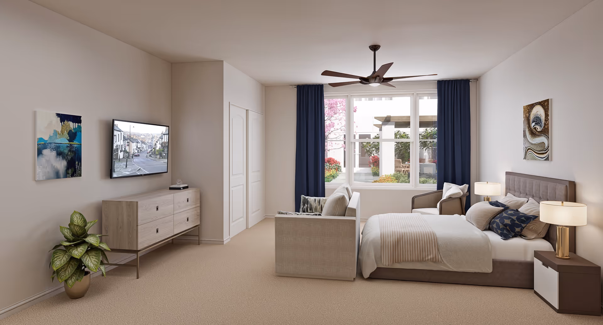 A bright and spacious bedroom with a large window showing a garden outside. The room features a bed with multiple pillows and a beige blanket, two bedside tables with lamps, a beige sofa at the foot of the bed, a single armchair, a ceiling fan, a wall-mounted TV above a wooden dresser, two pieces of wall art, and a potted plant on the floor.