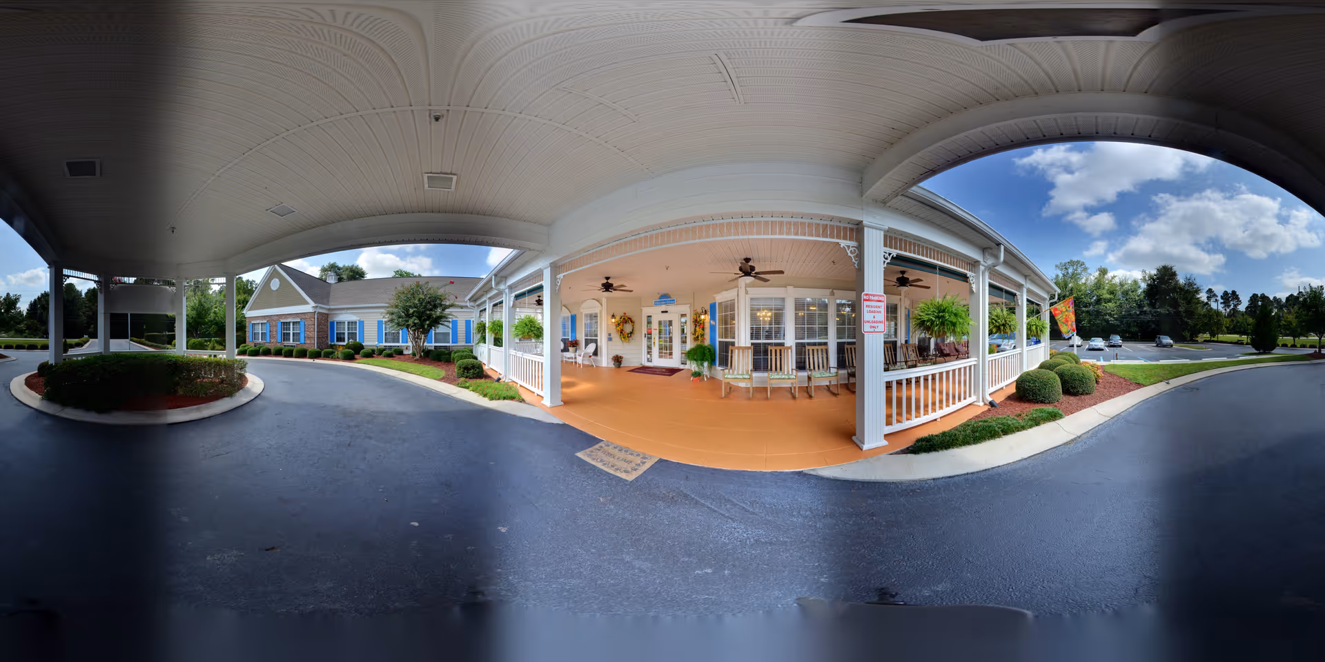 Covered driveway and entrance area of a senior living facility with white pillars, ceiling fans, rocking chairs, and hanging plants. The building has a brick and siding exterior with blue shutters, surrounded by landscaped bushes and trees under a partly cloudy sky.
